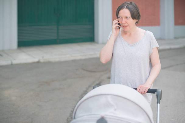 Upset and worried mother talking on mobile phone and pushing baby in a stroller while walking on the street in casual sport clothing - Fotoğraf, Görsel