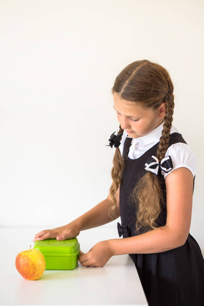 Koulupukuinen pikkutyttö kerää lounasta keittiössä koulua varten. Lapsi sulkee eväsrasian. Lounaslaatikon vieressä on omena. Poika on valmis kouluun. Takaisin kouluun.
. - Valokuva, kuva