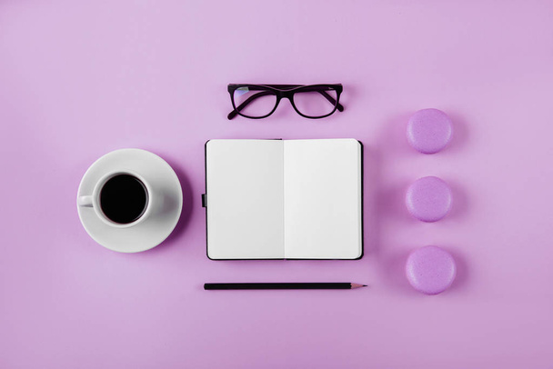 Coffee, macaron, clean notebook, eyeglasses and flower on violet table from above. Female working desk. Flat lay style. - Photo, Image