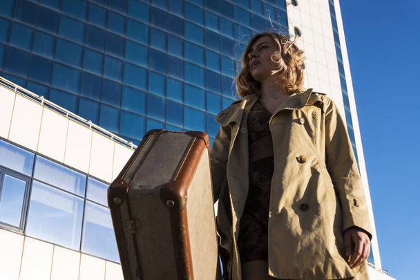 Attractive Young Blonde Woman in Trench walking in City with Vintage Suitcase - Fotó, kép