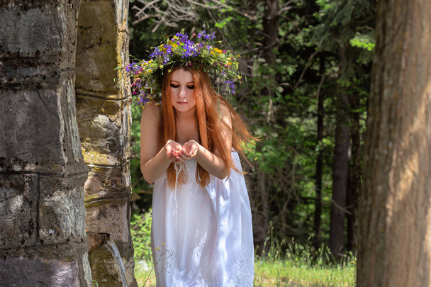 Joven, hermosa mujer en un vestido blanco y con una corona de flores silvestres en su cabeza cerca de una fuente en un bosque de montaña close-u
 - Foto, Imagen