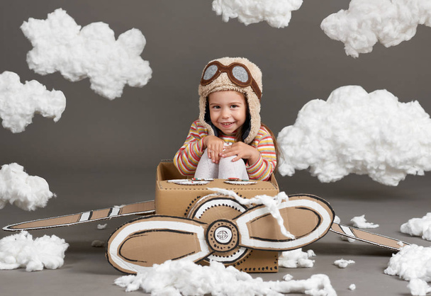 το παιδί κορίτσι παίζει σε ένα αεροπλάνο από χαρτόκουτο και ονειρεύεται να γίνει πιλότος, σύννεφα, βαμβάκι σε ένα γκρίζο φόντο - Φωτογραφία, εικόνα