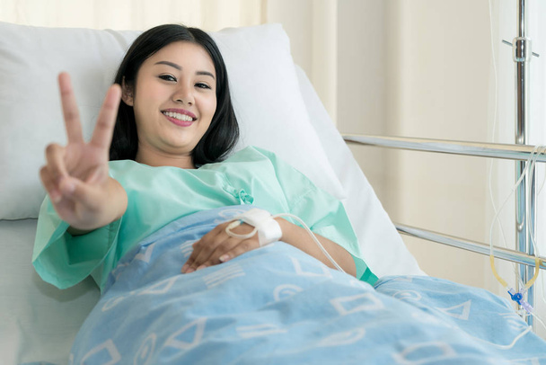 Genç Asyalı hasta kadın hastanede yatarken yalan neşeli kayıt zafer gösterilen tuzlu damla ile. Hasta yardımcı olmak için tuzlu çözüm damla. - Fotoğraf, Görsel