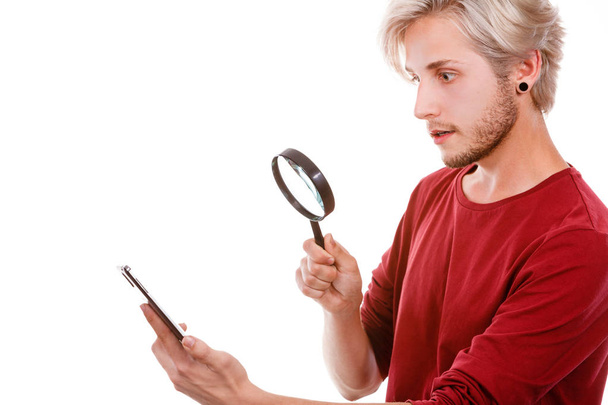 Funny young guy reading message on smart phone looking with ssurprised face expression, using loupe magnifying glass, isolated on white - Fotografie, Obrázek