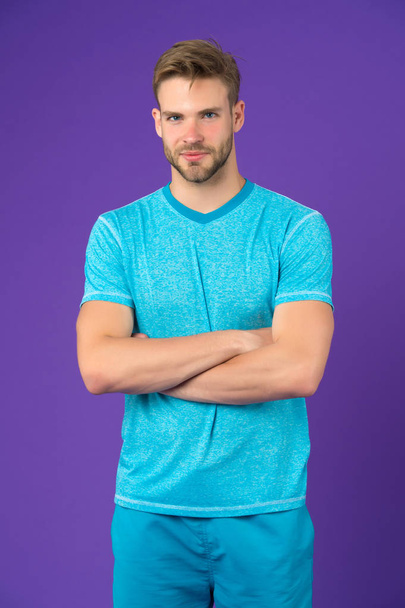 Man on smiling face posing confidently with folded arms, violet background. Guy with bristle wears set of sportive clothes . Fashion concept. Man looks attractive in sportive set of shirt and pants - Photo, image