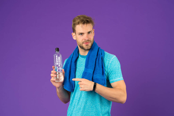 Menekşe arka plan üzerinde plastik şişe, sporcu noktası parmak. Adam tutun su şişesi. Mavi tshirt ve şort eğitimden sonra havlu ile atlet. Susuzluk ve dehidrasyon. Spor aktivitesi ve enerji - Fotoğraf, Görsel