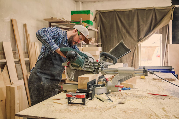 Experienced carpenter in work clothes and small buiness owner working in woodwork workshop, using a circular saw to cut through a wooden plank, on the table is a hammer and many tools - Photo, Image