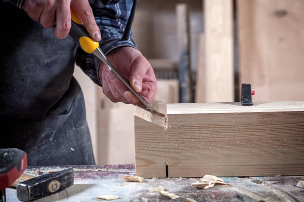 Εσωτερικη πεπειραμένος ξυλουργός σε ρούχα εργασίας και μικρά buiness ιδιοκτήτης εργάζεται σε εργαστήριο ξυλουργικής, χρησιμοποιώντας μια σμίλη για να αποκόψει από ξύλο σε εργαστήριο στο τραπέζι είναι ένα σφυρί και πολλά εργαλεία  - Φωτογραφία, εικόνα