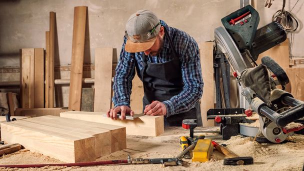 carpintero experimentado en ropa de trabajo y pequeño propietario buiness mide una tabla de madera con una regla y marcas con lápiz los puntos necesarios para rebanadas
 - Foto, Imagen