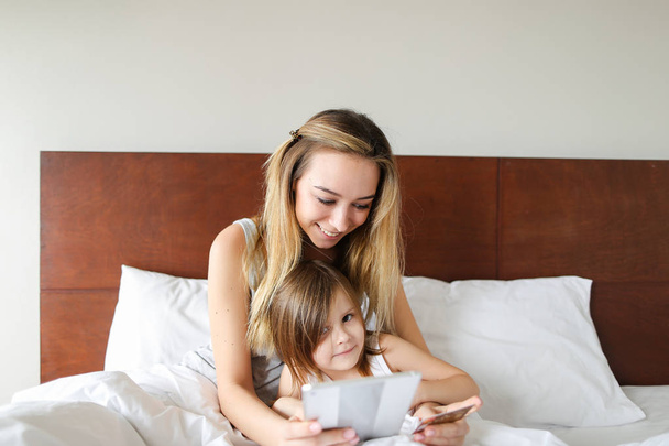 Genç beyaz anne ile küçük kızı yatakta oturuyor ve tablet kullanma. - Fotoğraf, Görsel