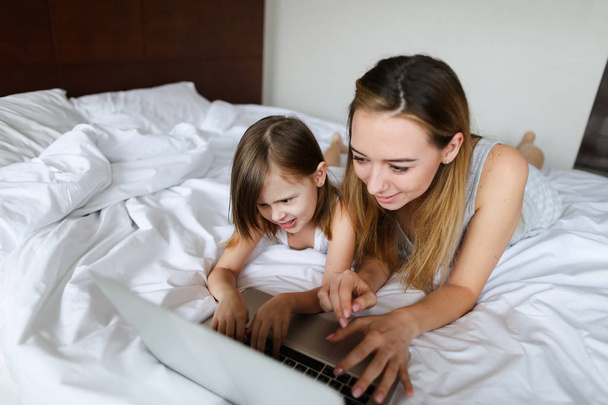 Nuori kaunis nainen, joka käyttää kannettavaa tietokonetta pienellä tyttärellä ja makaa sängyssä
. - Valokuva, kuva