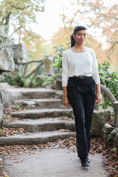 Leuk zwart meisje dragen witte blouse staande in de buurt van stenen trap in park. - Foto, afbeelding