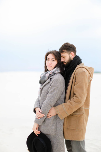 Kaukasische jongeman knuffelen vrouw grijze jas en sjaal dragen in witte winter achtergrond. Concept van romantische paar fotosessie en seizoensgebonden inspiratie. - Foto, afbeelding