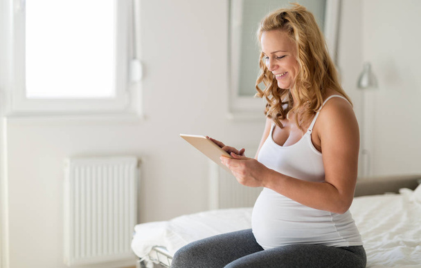 Happy pregnant blonde woman using tablet in bedroom - Fotó, kép