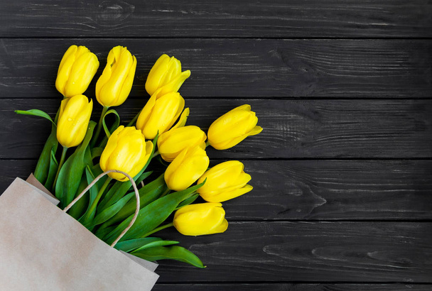 Bright yellow tulips in eco paper bag on black vintage wooden table. Flat lay, top view. Spring, shopping, eco concept - Zdjęcie, obraz