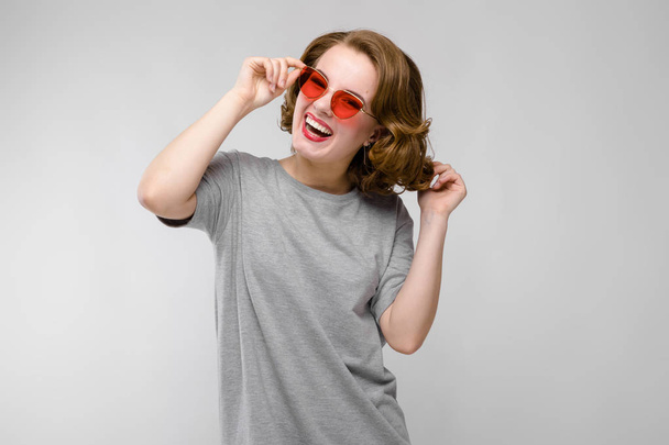 Charming young girl in a gray T-shirt on a gray background. Happy girl in red glasses. The girl laughs - Photo, Image