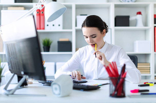 Una joven en la oficina tiene un bolígrafo en la boca y trabaja con una calculadora, documentos y una computadora
. - Foto, Imagen