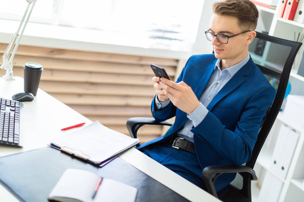 Um jovem está sentado com um telefone em suas mãos em uma mesa no escritório
. - Foto, Imagem