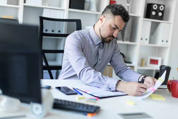 Молодий чоловік в офісі сидить за столом, тримає маркер в руці і працює з документами
. - Фото, зображення
