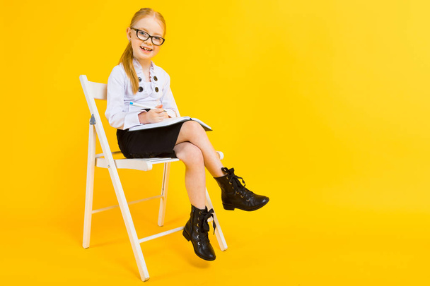 Girl with red hair on a yellow background. A charming girl in transparent glasses sits on a white chair and makes notes in a notebook. - Foto, afbeelding