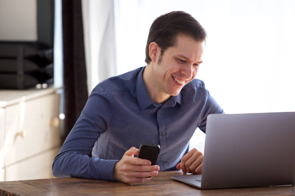 Detailní záběr portrét pohledný usmívající se muž pracuje na notebooku s mobil - Fotografie, Obrázek