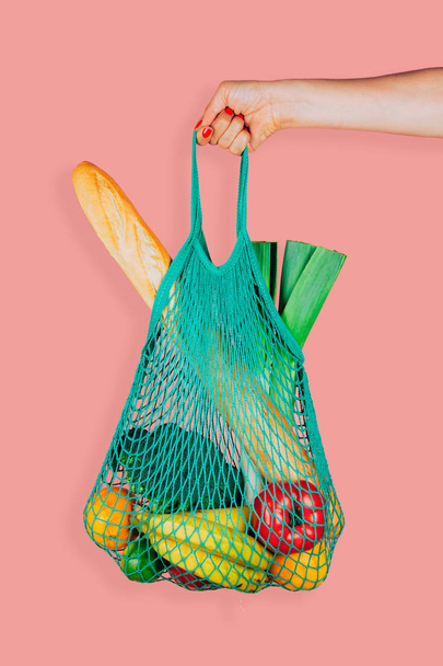 Woman hand holding a green mint string shopping bag with vegetables, fruits and bread in front of a pink background - 写真・画像
