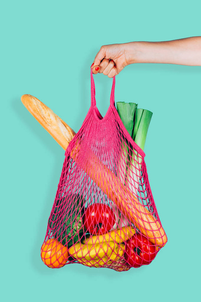 Woman hand holding a fuchsia string shopping bag with vegetables, fruits and bread in front of a green mint background - Photo, Image