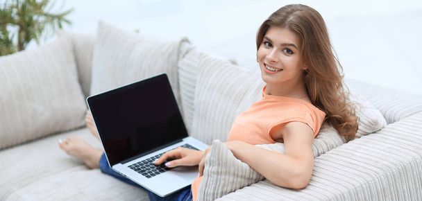 美しい若い女性のソファーに座っているとカメラを見てはノート パソコンでの作業. - 写真・画像