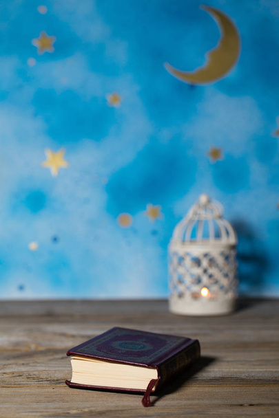Священная книга Корана на деревянной поверхности. Голубое ночное небо с полумесяцем и подсвечником на заднем плане
 - Фото, изображение