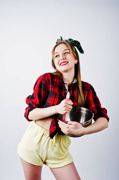 Young funny housewife in checkered shirt and yellow shorts pin up style with saucepan and kitchen spoon isolated on white background. - Foto, afbeelding