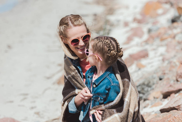 happy mother and daughter in sunglasses with blanket sitting on shore - Photo, image