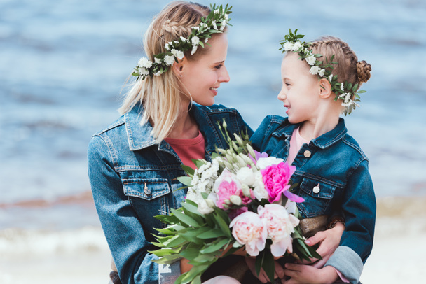 mother and daughter in floral wreaths with peony bouquet looking at each other on sea shore - Foto, imagen