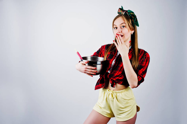 Young funny housewife in checkered shirt and yellow shorts pin up style with saucepan and kitchen spoon isolated on white background. - Photo, Image