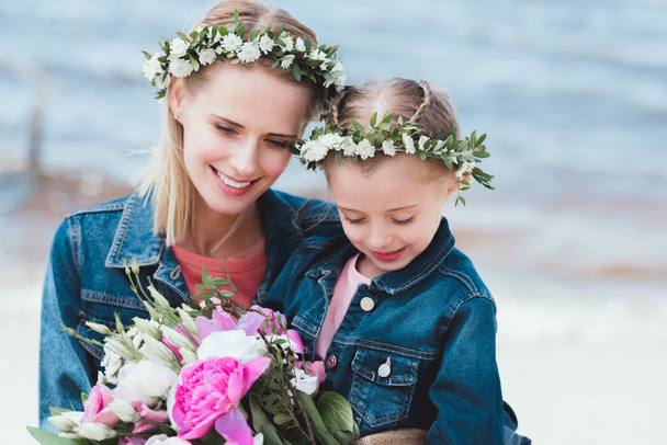 beautiful mother and smiling daughter in floral wreaths with bouquet on sea shore - Foto, imagen