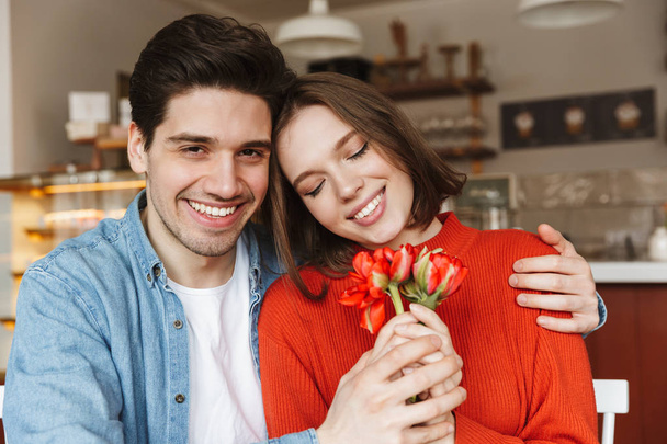 Fénykép fiatal szerető pár férfi és a nő gyönyörű virágok összetartja, hangulatos kávézó asztalnál ülve - Fotó, kép