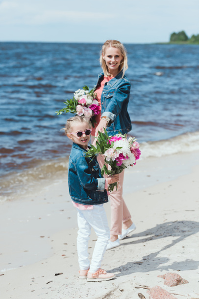 mother and daughter with peony bouquets holding hands on sea shore - Photo, Image