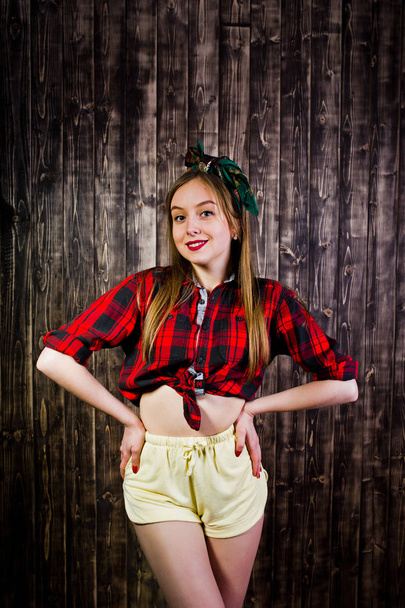Young funny housewife in checkered shirt and yellow shorts pin up style on wooden background. - Фото, зображення