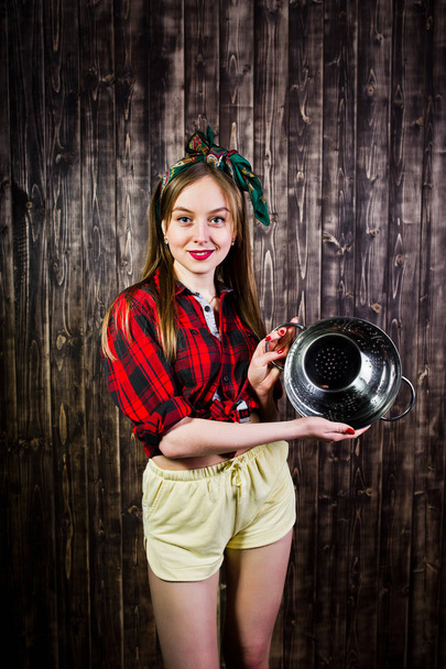 Young funny housewife in checkered shirt and yellow shorts pin up style with kitchen sieve on wooden background. - Foto, afbeelding
