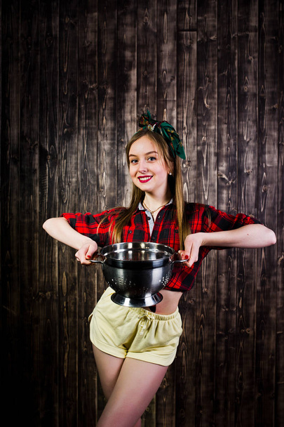Young funny housewife in checkered shirt and yellow shorts pin up style with kitchen sieve on wooden background. - Valokuva, kuva