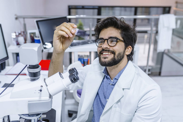 Life scientist researching in laboratory. Scientist examines biopsy samples. Satisfied male researcher smiling cheerfully while sitting at a microscope and examining a sample in his hands. - Фото, изображение