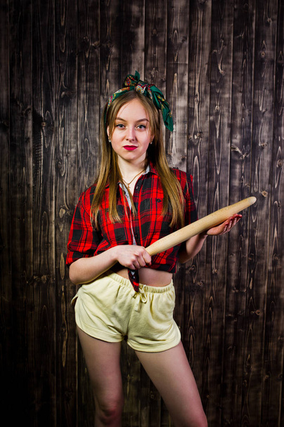 Young funny housewife in checkered shirt and yellow shorts pin up style with kitchen rolling pin on wooden background. - 写真・画像