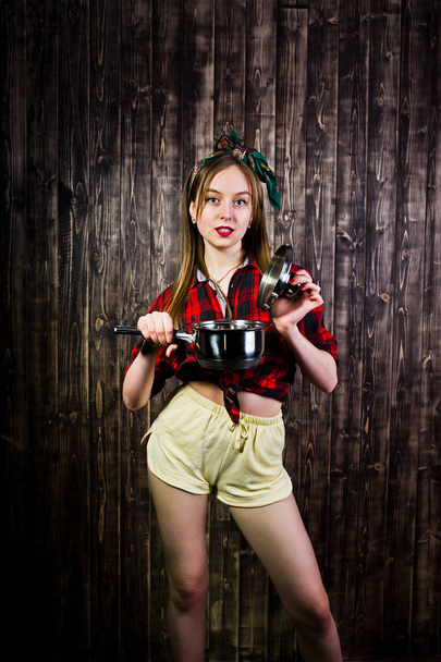 Young funny housewife in checkered shirt and yellow shorts pin up style with saucepan on wooden background. - Фото, изображение