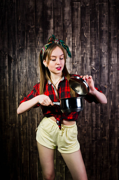 Young funny housewife in checkered shirt and yellow shorts pin up style with saucepan on wooden background. - Zdjęcie, obraz
