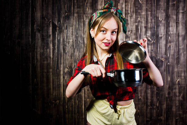 Young funny housewife in checkered shirt and yellow shorts pin up style with saucepan on wooden background. - Valokuva, kuva