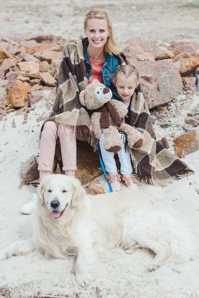 attractive mother and daughter in blanket sitting with dog on shore - Photo, Image