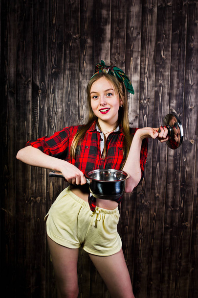 Young funny housewife in checkered shirt and yellow shorts pin up style with saucepan on wooden background. - Foto, Bild