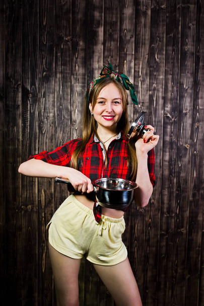 Young funny housewife in checkered shirt and yellow shorts pin up style with saucepan on wooden background. - Photo, image