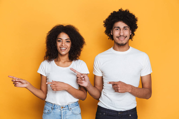 Portrait of a cheerful young afro american couple pointing fingers away at copy space isolated over yellow background - Foto, immagini