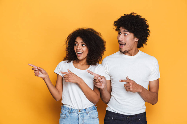 Portrait of a happy young afro american couple pointing fingers away at copy space isolated over yellow background - Fotografie, Obrázek