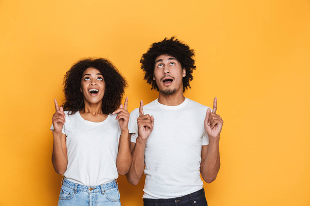 Portrait of a happy young afro american couple pointing fingers up at copy space isolated over yellow background - Photo, Image